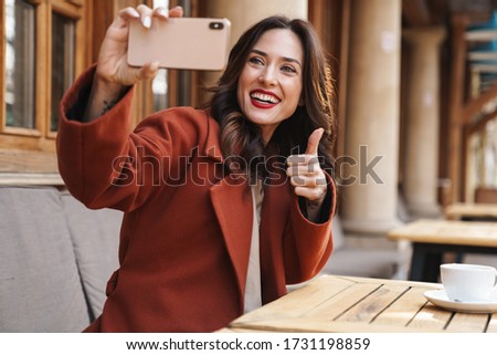 Image of smiling adult woman taking selfie on cellphone and showing thumb up while sitting in cafe outdoors