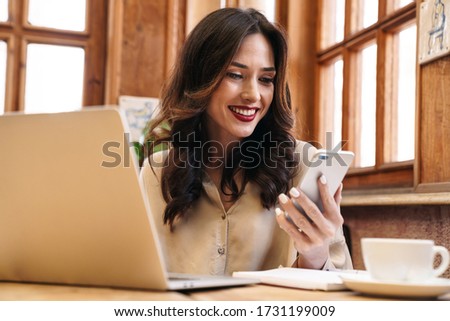 Image of happy brunette adult woman using cellphone and working with laptop while sitting in cozy cafe indoors