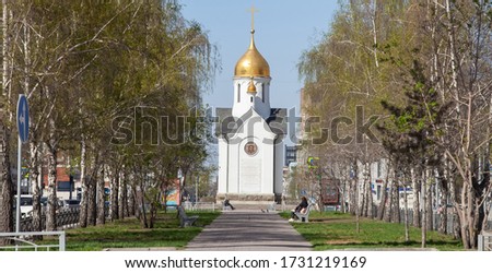Chapel named after St. Nicholas the Wonderworker in Russian city of Novosibirsk is white with a golden dome next to a bench in an alley of birches located between two roads with different directions.