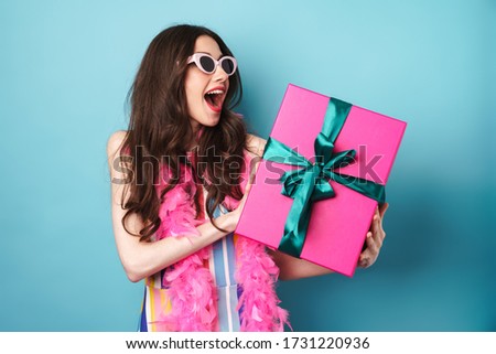 Image of surprised brunette woman in sunglasses holding gift box isolated over blue background
