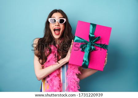 Image of surprised brunette woman in sunglasses holding gift box isolated over blue background