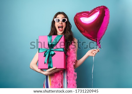 Image of surprised brunette woman in sunglasses holding gift box and balloon isolated over blue background