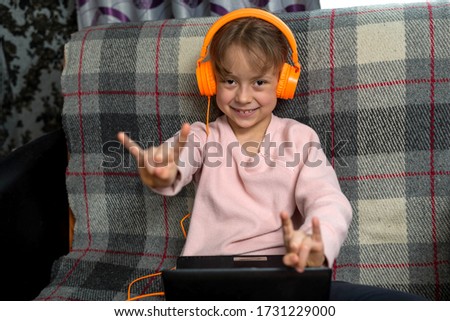 Cute little girl sitting in headphones and with a laptop on the sofa in the room. The concept of distance learning during quarantine.