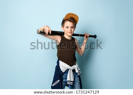 Naughty kid looking forward with a sly smile on face keeping baseball bat behind his head. Adolescent, active, ruffian, mischievous child. Three quarter length portrait isolated on blue background