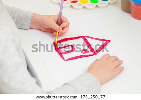 A child's hand with brush is drawing picture of country house by watercolor on white paper. Side view.