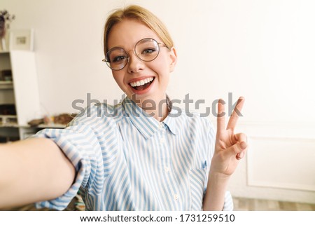 Photo of joyful blonde woman in eyeglasses showing peace sign while taking selfie photo at office