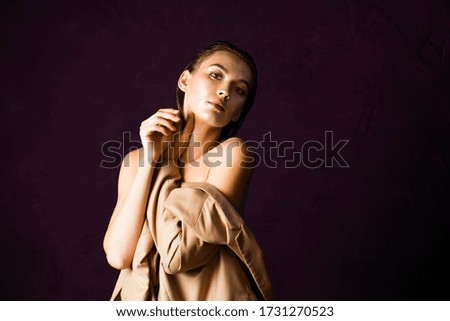 Woman with natural make up in a beige jacket put on on hand is siting on a chair. Photography with dark magenta and orange-gold-beige tones. High contrast and dark background with a copyspace