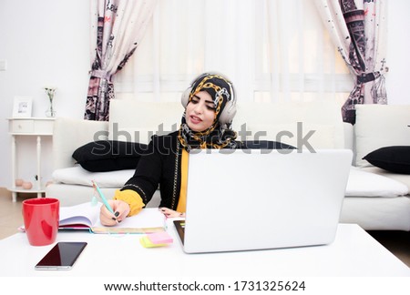 Arabic woman working from home and taking notes on a notebook under a call meeting using headphone and wearing abaya and hijab.