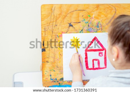 Child is drawing yellow sun with red house on white sheet of paper of easel during education at school. Children creativity.