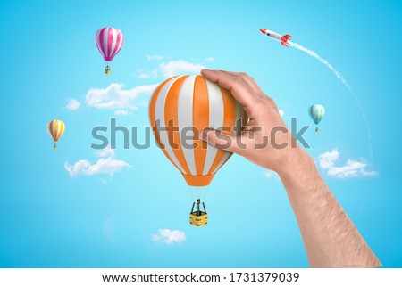 Hand holding orange white hot air balloon with silver rocket and hot air balloons on blue sky background. Concept art. Flying objects. People and objects.