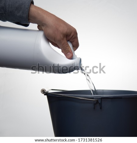 A man's hand pouring cleaner or bleach into a blue bucket against a white background. Royalty-Free Stock Photo #1731381526