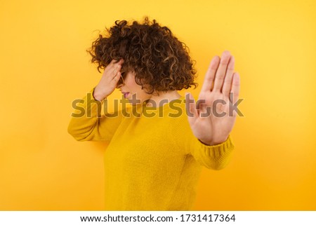 People, body language. Young woman covers eyes with palm and doing stop gesture, tries to hide from everybody. Don't look at me, I don't want to see, feels ashamed or scared. Wearing yellow sweater