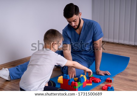 Young male doctor with beard in a blue uniform sitting on the floor next to the boy 10 years, they play educational toys, concept rehabilitation.