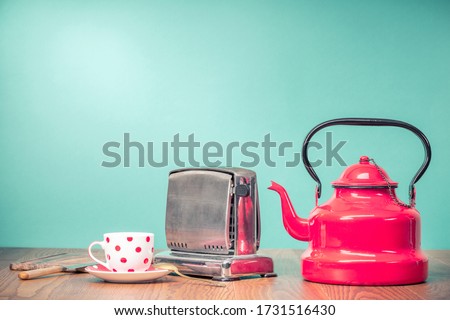 Retro classic red kettle, a cup of tea, outdated bread toaster, kitchen board and vintage knife on oak wooden table in front mint green background. Nostalgic equipment. Old style filtered photo