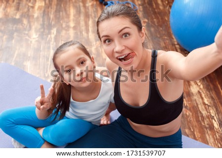 Young woman and her charming daughter sitting together at home, making funny faces for selfie portrait, showing victory sign and smiling nice