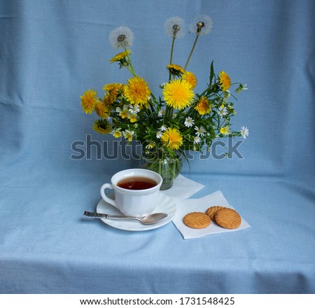 Still life on a blue background with a bouquet of spring flowers of bright yellow dandelions, with a white cup of tea, and cookies located on the table, the concept of a spring morning in delicate sha