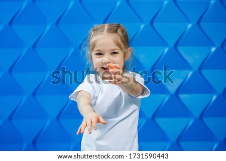 A girl with blond hair in a white T-shirt on a blue background. She smiles and waves her hands