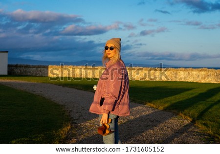 Girl with camera wearing sunglasses hat and pink jacket standing on stone path