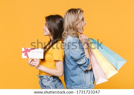 Side view of young women girls friends in denim clothes isolated on yellow background. People lifestyle concept. Hold package bag with purchases after shopping gift certificate, standing back to back