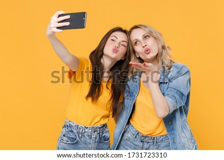 Two pretty young women girls friends in casual t-shirts denim clothes posing isolated on yellow background studio. People lifestyle concept. Doing selfie shot on mobile phone blowing sending air kiss