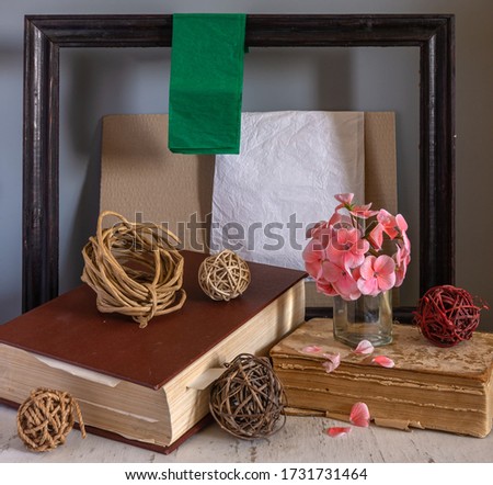 On the table are books and tangles of ropes and branches. Blooming geranium. Wooden frame for paintings and sheets of paper. Interior.
