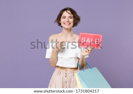 Smiling young woman in summer clothes isolated on violet background. Shopping discount sale concept. Mock up copy space. Hold package bag with purchases pointing index finger on sign with SALE title