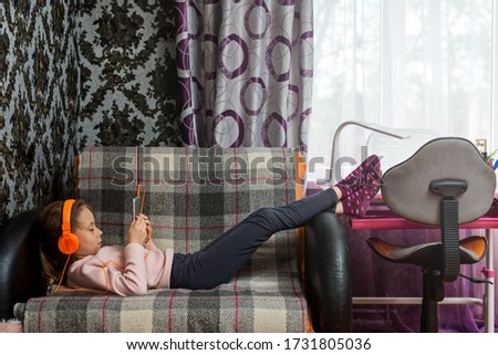 Cute little girl lies in headphones and with a tablet on the sofa in the room. The concept of distance learning during quarantine.