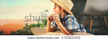 Boy in straw hat lying down, imitating scouting with field-glass at home near TV screen. Funny hiking, camping. Family time. Crazy activities. Coronavirus situation. Quarantine. Isolation from covid19