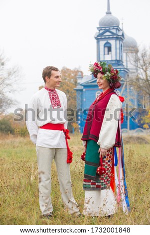 Girl and guy in retro costumes on the street in the old village. Retro staging of an ancient rite. Beautiful wreath on a girl. The guy hugs the girl, both are smiling. Antique clothing of the late