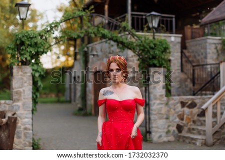 Portrait of attractive redhead tattooed woman in red dress and diadema on her head posing on blurred medieval castle background at summer sunset