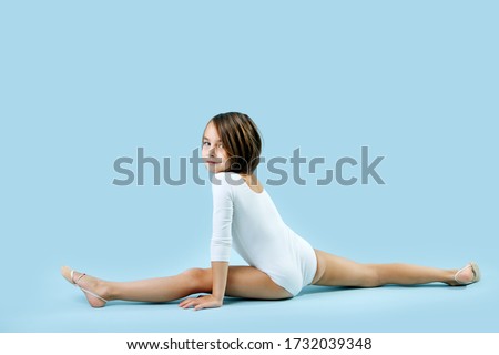 Little brunette girl in a white leotard sitting in front splits, smiling at camera over blue background.