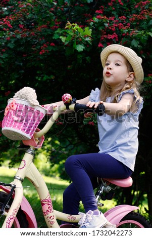 Little happy girl sitting on a bicycle and playing. She is wearing a hat and a T-shirt. Summer warm day. Flowering bush on a background