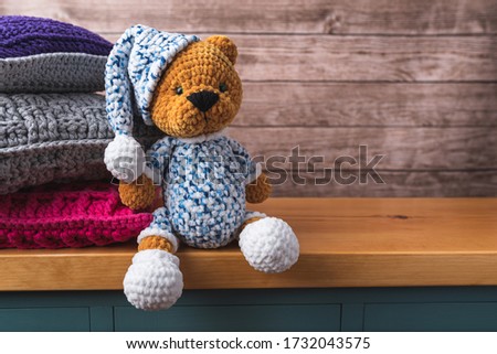 Brown little bear with hat made of yarn. Plush toy sitting on a cabinet. Pillows in the background. Handiwork. Composition with free copy space for text or graphics. Teddy bear.