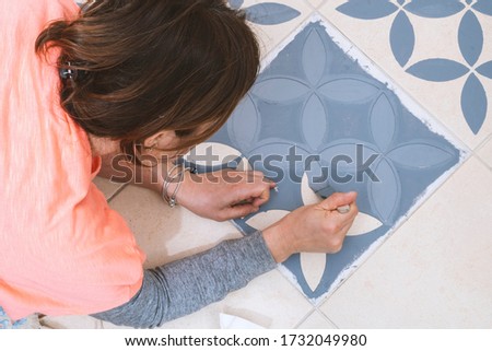 Stay at home and home improvement concept: Close up and top view of a woman holding a brush to paint a decorative template on the floor tiles into gray by using a generic turkish pattern stencil Royalty-Free Stock Photo #1732049980