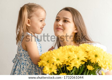 motherhood, mothers day, birthday, childhood, family concept - close-up little girl in dress congratulate and give young mother bouquet of yellow daisy flowers and kisses her on white background.