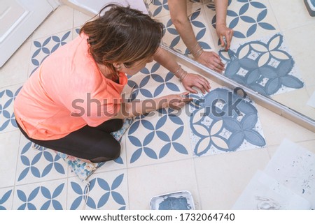 Stay at home and home improvement concept: Top view of a woman painting with a brush a decorative template on floor tiles in gray by using a generic turkish pattern stencil. Reflection in mirror Royalty-Free Stock Photo #1732064740