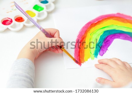 Top view of hands of child drawing a rainbow with brushes and paints on white paper.