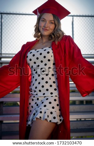 A cute girl in a polka dot dress and cap and gown stands on bleachers and poses for a graduation portrait picture.
