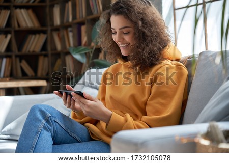 Happy young hispanic latin teen girl sit on sofa at home holding phone looking at screen watching social media video content, movie or stream, video calling online in mobile app using smart phone. Royalty-Free Stock Photo #1732105078