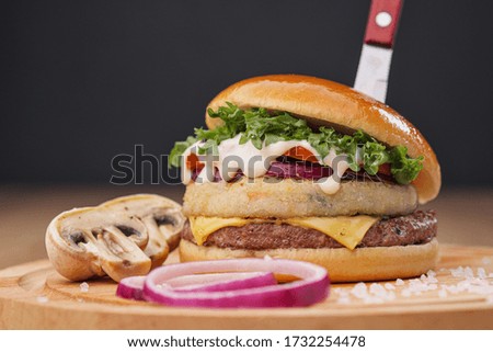 Delicious juicy hamburger on a served wooden board with a stuck knife, on a black background. Cutlets, buns, mushrooms and vegetables. Mushroom hamburger.