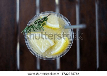 Lemonade with ice. Pieces of lemon with ice decorate by rosemary. Famous drink in summer. Top table picture with wood table background.