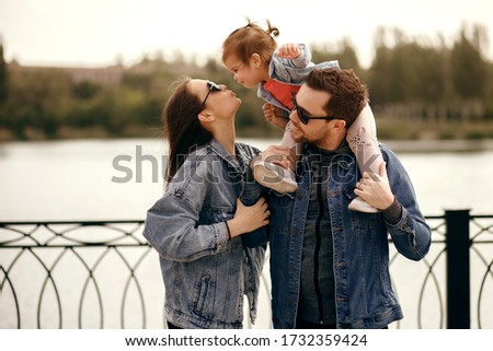 little daughter sits on dad shoulders and kisses mom, fashionable parents are dressed in jeans clothes, sunglasses, a beautiful young family walks along the river embankment and enjoys at sunset