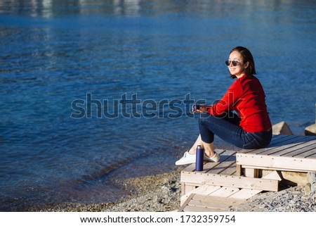 Happy young woman smiles joyfully sitting on seashore on wooden steps with her legs crossed and drinks tea or coffee in evening on background of water. Life after quarantine. Closer to nature.