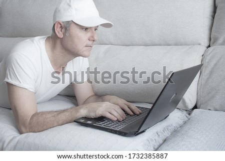 Middle-aged man working with laptop on the couch at home, wearing white casual shirt and cap.  Horizontal photography in white color