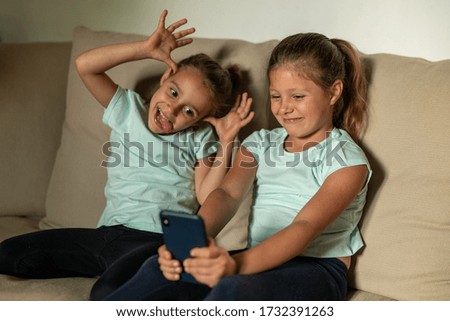 Authentic shot of two happy little sisters are having fun to make a selfie or video call with a smartphone to parents or relatives while sitting on a sofa in a living room at home.