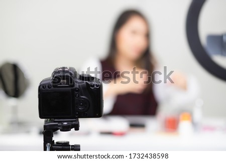 The DSLR digital camera is mounted on a tripod, taking pictures or shooting videos in the room with a blurry background. A woman is selling, picking up cosmetics in a live broadcasting 