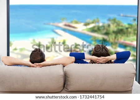 Rear view of a couple relaxing on a sofa at hotel and looking outside a sea background through the window of the living room
