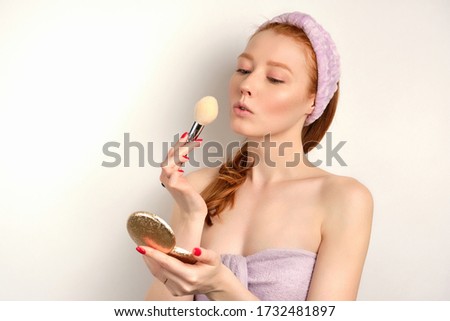 Cute red-haired girl stands on a white background in a towel with a brush in her hands and looks in the mirror, focus on the face