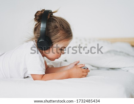 Cute toddler girl with fair hair in headphones with mobile on bed in bright interior