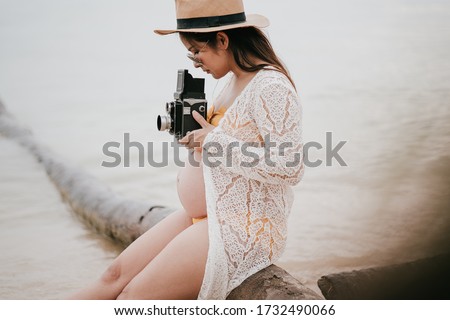 Beautiful asian young pregnant woman wearing orange bikini is  sitting to take a picture of the sunset by the sea at Phangan island in evening time. Selective focus and split color.